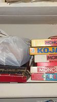 Shelf with stacked vintage board game boxes including Monopoly, Kojak, and Careers, plus a plastic bag of game pieces.