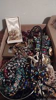 Photo showing a variety of costume jewelry including beaded necklaces, bracelets, and brooches in multiple colors and styles inside a cardboard box.