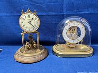 Two Vandor clocks, one tall pendulum style and one smaller glass dome clock on a brass base