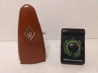 Photo showing both metronomes side by side on a white surface: brown Taktell Piccolo and black Seiko Quartz metronome