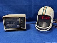 Photo showing white helmet-shaped radio with red LED display indicating time 6:54 alongside beige Emerson digital clock radio with flip numbers showing 11:21.