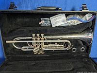 Overall view of the silver trumpet inside its black hard case with accessories stored in the case compartments.