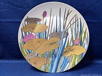 Front view of a large round Rosenthal decorative dish showing detailed botanical design with lily pads, flowers, and reeds in earth tones and blue-green shades on a white base.