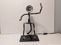 Full frontal view of adjustable metal robot lamp on black rectangular base showing articulated arms, legs, and exposed joints with power cord and switch visible behind.