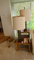 Two tall lamps with beige fabric shades sitting on a wooden accent table near window with rolled posters beside them