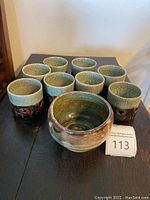 Set of 8 matching handmade pottery cups and 1 bowl, displayed on a black table. Cups have a light blue-green crackled glaze top and brown textured lower half.