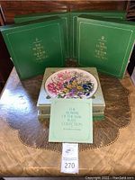 Full set of 12 Franklin Porcelain green boxes stacked and one plate displayed on top along with an accompanying information leaflet titled 'The Flowers of the Year Plate Collection'