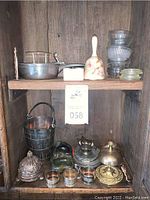 Overview of items on wooden shelf including pewter bucket, napkin rings, footed dishes, brass bell, porcelain bell, glass bowls, pewter pot, and wire basket