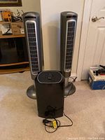 Two silver Lasko tower fans with an Oreck fan in front on carpeted floor.