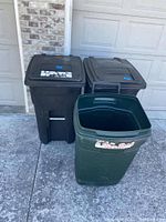 Three outside trash cans: two black wheeled with lids and one green open top, placed on driveway in front of garage door.