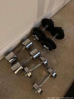 Photo showing six dumbbells arranged on carpet against a wall; includes two black coated 20 lb dumbbells and four chrome plated dumbbells.