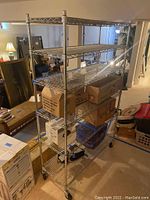 Full view of the five shelf metal wire rack with caster wheels. Several boxes and items stored on middle shelves for scale and perspective.