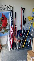 Wide view of garden tools including shovels, rakes, brooms, flags on poles and metal flag stand with cardinal flag