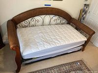 Sofa bed with visible wooden armrests and wicker sides, metal scrollwork backrest, twin mattress on top, and pullout frame underneath