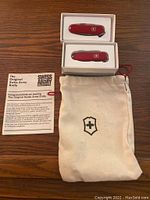 Two red Victorinox Swiss Army knives in white boxes, a beige carrying pouch, and an informational card laid out on a wooden surface.