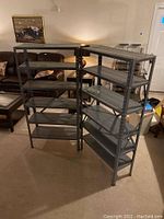 Two gray metal shelving units standing side by side indoors on carpeted floor with living room furniture in the background. Each unit has 5 shelves.