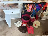 Wide view of floor showing several wigs stored in containers, multiple colorful scarves draped on drawer, white gloves, beige sheep hat, and boxed hosiery.