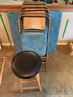 Four bronze-tone metal folding chairs stacked with blue padded seats and backrests, alongside a matching padded folding card table. Wooden stool with padded black seat is in front.