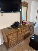 Front view of wood dresser with multiple drawers and geometric-patterned cabinet doors, topped with various items and mounted mirror above.