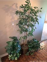 Image showing three artificial plants positioned against a wall with visible light shadows, including one tall faux tree and two smaller faux greenery plants.