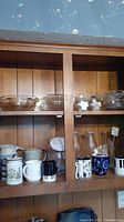 Wooden shelving with various clear glass bowls, ceramic canisters and mugs, and clear glass pitchers and bottles arranged on shelves.