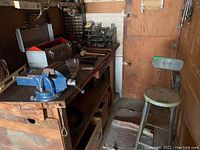 Wide view of wooden workbench with blue metal vise, multiple hammers and mallets, and metal shop stool