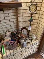 Photo showing black metal clock on scroll-design stand, brown wooden round clock, pink faux floral tulip bouquet, white swan figurines, pink/brown basket with bows, and a small silver metal container arranged on a brick hearth.