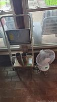 Foldable metal step ladder, small wooden stool with two metal cups on top, and white oscillating table fan all placed against a glass window.