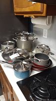 Photo shows a collection of various pots and pans including frying pans, saucepans, soup pots, steamer pot, all with metal handles and several with lids.