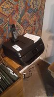 Photo showing black HP OfficeJet 4655 printer with phone handset on top, placed on wooden table near patterned wall.