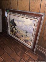 Photo of the framed oil painting resting against a wood-paneled wall showing the ornate frame and partial view of the artwork.