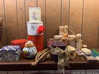 Full view of various decorative tin canisters alongside pillar candles and a doll-shaped container on a wooden surface against wood-paneled wall.
