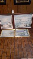 Four framed landscape paintings on the floor with wood and gold frames, featuring lighthouses and coastal scenes