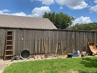 Wide view of wooden ladder, metal galvanized tub, various long-handled yard tools including shovels, rakes, hoes, garden forks, rusty wheelbarrow, and an enamel pot against a wooden fence in backyard.