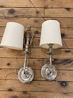 Top view of both wall sconces showing polished nickel finish and white fabric shades, placed on wooden floor.