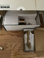 Partially open Kohler box showing brass hardware pieces for wall sconce and separate box containing toilet paper holder parts on wooden surface.