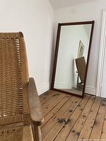 Full view of large floor mirror with teak wood frame standing against wall beside rustic wooden chair on wooden floor.