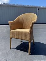 Front angle view of Lloyd Loom wicker chair outdoors on pavement, showing woven natural finish, curved backrest and arms, and four legs.