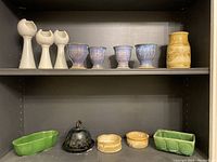 Various pieces of studio pottery including green planters, blueish cups, butter dish, and other ceramic items arranged on black shelving.