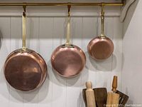 Exteriors of three copper frying pans hanging on brass rail