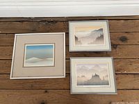 Three framed and matted Markgraf prints labeled Morning Sun, Evening Sky, Drifting Snow on a wooden floor showing full view of all three artworks and frames.