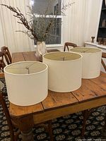 Three cream-colored drum lampshades displayed on a wooden table with chairs and decorative dried flowers in the background.