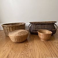 Four vintage woven baskets arranged on wood floor, including a large oval light brown basket, a large dark brown picnic basket with handles, and two smaller round baskets, one with lid.