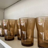 Close-up side view of 6 large amber glass tumblers on white shelf.