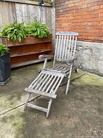 Full view of weathered teak steamer chair with extended footrest placed outdoors on concrete next to brick and stone wall.