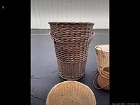Full height wicker laundry basket standing upright on asphalt ground with other baskets in the background.