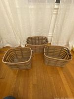 Three wooden rectangular baskets with leather handles positioned on wooden floor near white curtains, showing all baskets together.