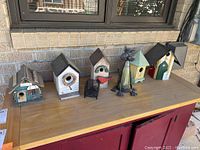 Photo of five wooden bird houses arranged on a table outside, with the metal cat sculpture and the small candle holder between them.