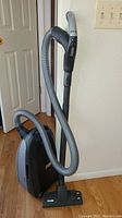 Full view of Miele canister vacuum with hose and floor nozzle standing upright indoors on wooden floor.