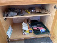 Photo of tools and painting supplies stored on cabinet shelves including plastic sheeting, rollers, painter's tape, putty knife, and sanding sponges.
